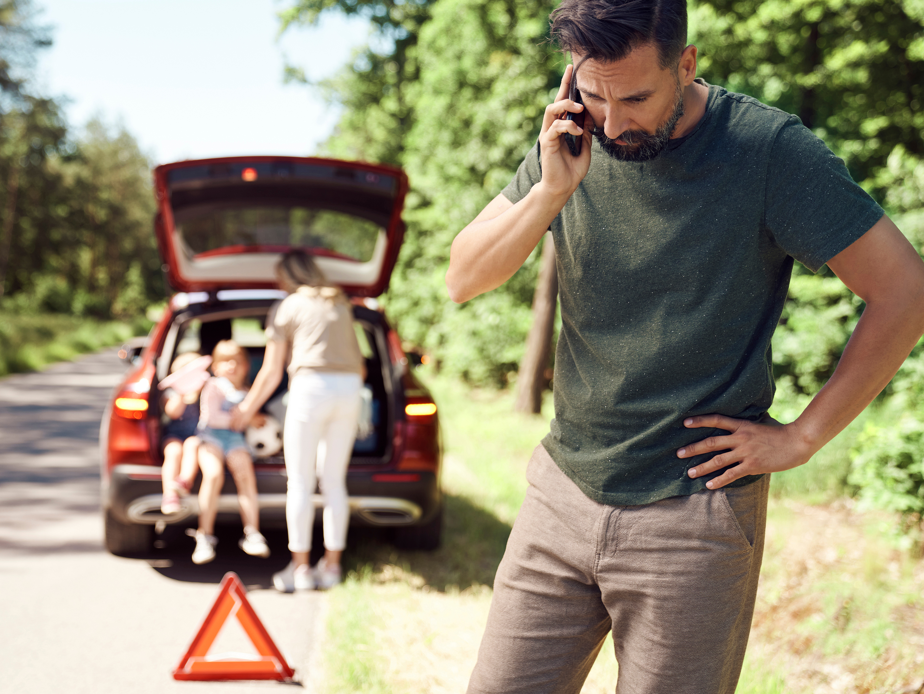 Man standing on side of road on phone, family in back of car